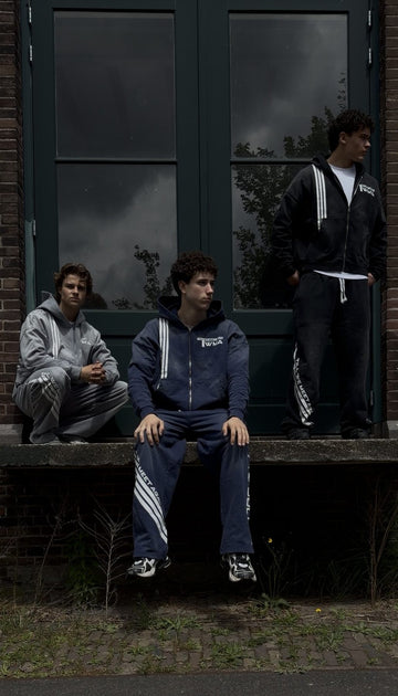 Drie modellen in de TWMA Faith Over Fear '26 trainingspakken in grijs, navy en zwart voor een groene deur