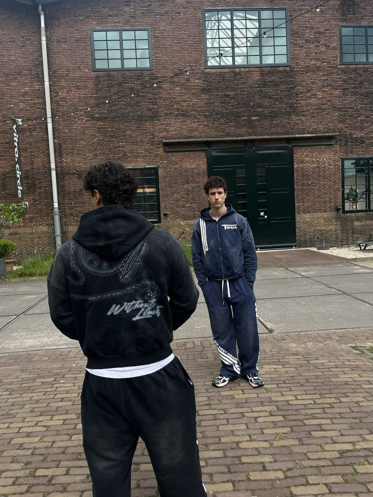 Twee modellen in de TWMA Faith Over Fear '26 zip-up hoodie en jogger in zwart en navy voor een bakstenen gebouwhero-slide