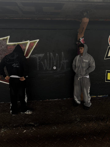 Twee modellen in zwarte en grijze TWMA-trainingspakken voor een muur met TWMA-graffiti