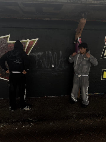 Model in zwarte TWMA-hoodie met 'Without Limits'-rugprint en model in grijs trainingspak bij een graffitimuur