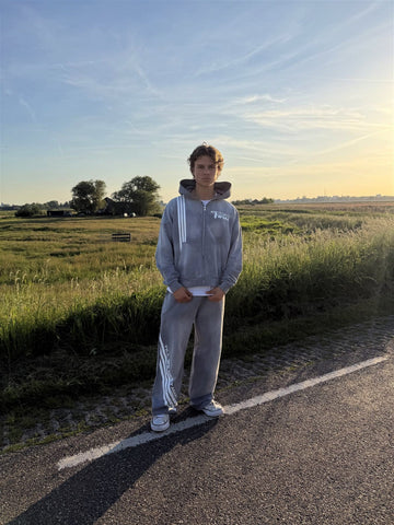 Model in het grijze TWMA Faith Over Fear '26 trainingspak op een polderweg bij zonsondergang