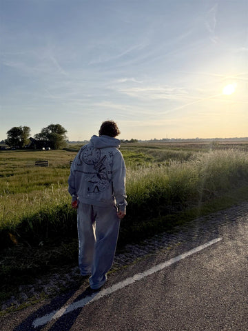 Model loopt over een polderweg bij zonsondergang in de grijze TWMA-hoodie met klokprint op de rug