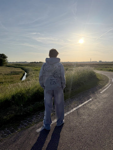 Model kijkt naar de zonsondergang in de grijze TWMA-hoodie met klokprint op de rug